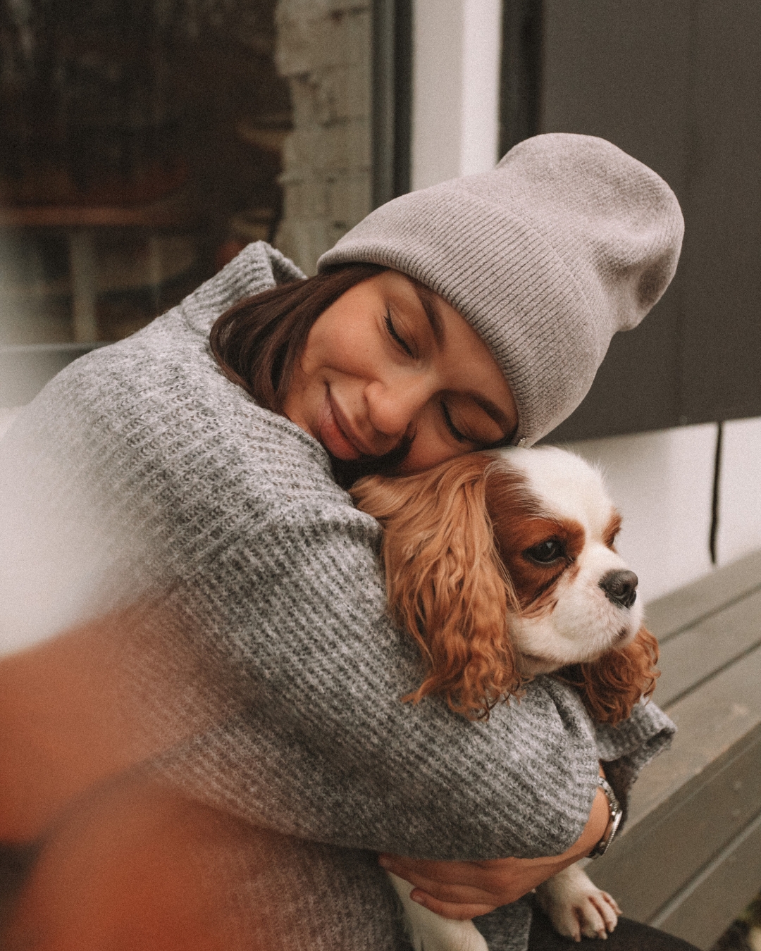 A woman hugging a brown and white dog while sitting on a bench, wearing a gray beanie and sweater