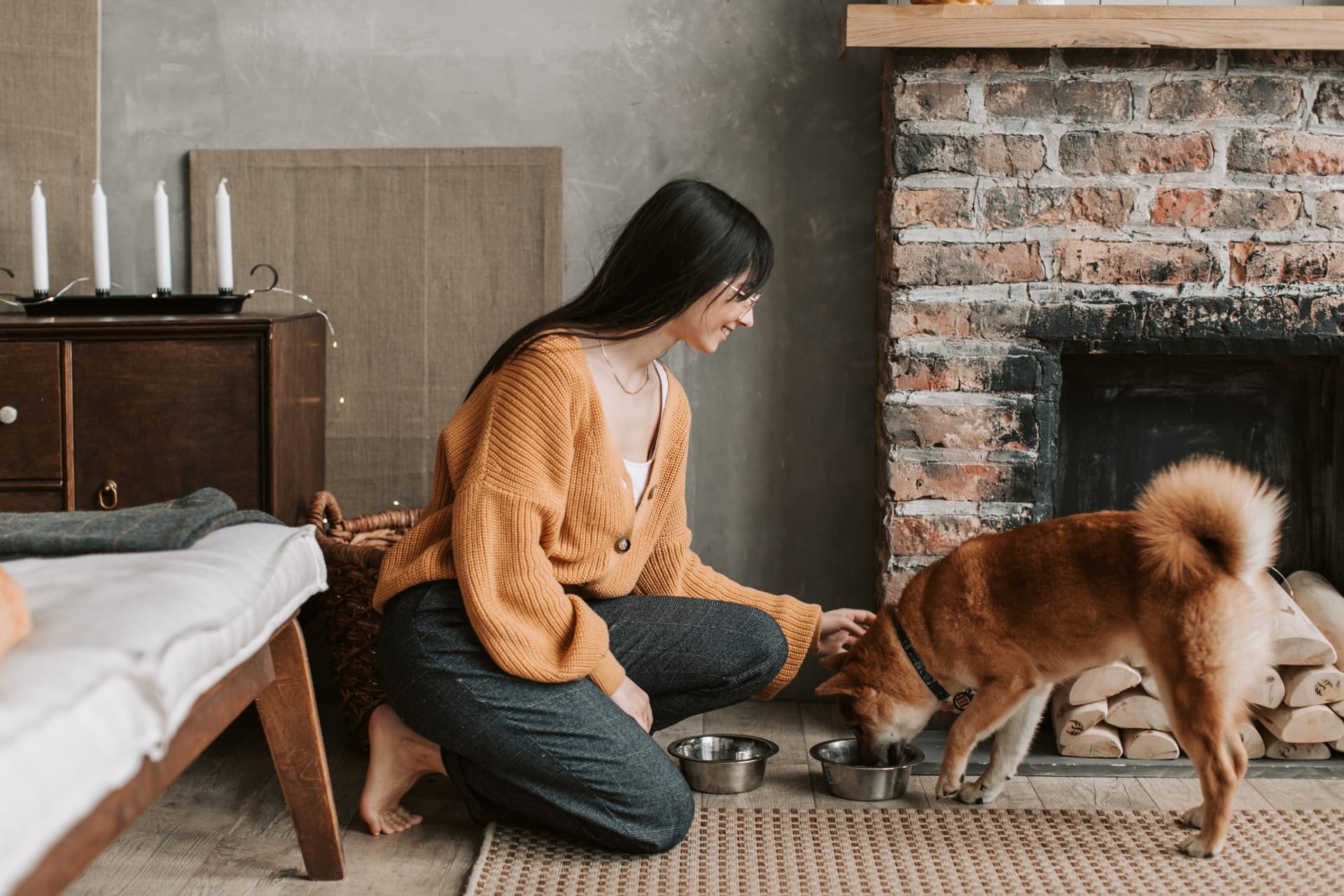 A woman with long dark hair, wearing an orange cardigan and dark pants, kneels on a textured rug in a cozy room with a brick fireplace. She is gently petting a Shiba Inu dog with a curled tail, which is eating from a metal bowl. The room features a wooden daybed with a blanket, a wicker basket, and a side table with white candles and string lights, creating a warm and inviting atmosphere. Stacked firewood is visible near the fireplace in the background.