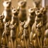 A collection of golden cat statues, styled in an ancient Egyptian design, is arranged closely together against a warm, blurred background. The statues vary in size, with some adorned with decorative collars and earrings, depicting cats in a seated, upright position with alert ears and expressive faces.