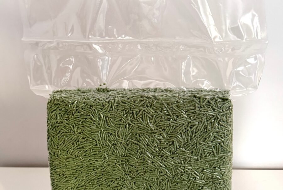 A vacuum-sealed plastic bag filled with green, cylindrical cat litter pellets, placed on a white surface against a light gray background.