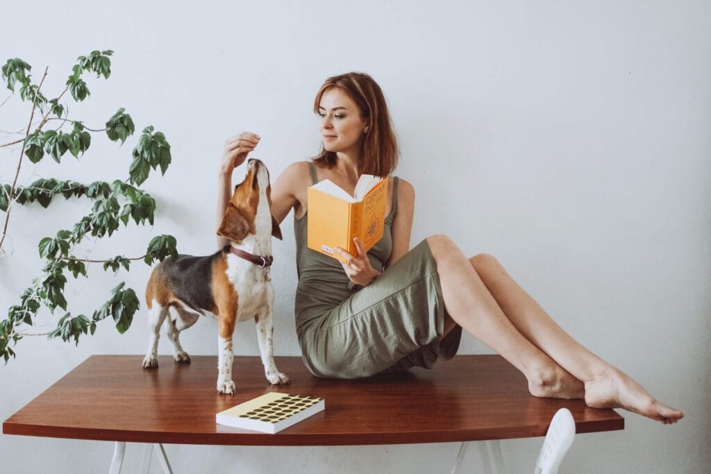 Una donna in un vestito verde è seduta su un tavolo di legno, legge un libro arancione, mentre un piccolo cane le sta accanto, guardando in alto verso un biscotto che lei tiene in mano. Una pianta verde e un quaderno sono nelle vicinanze.