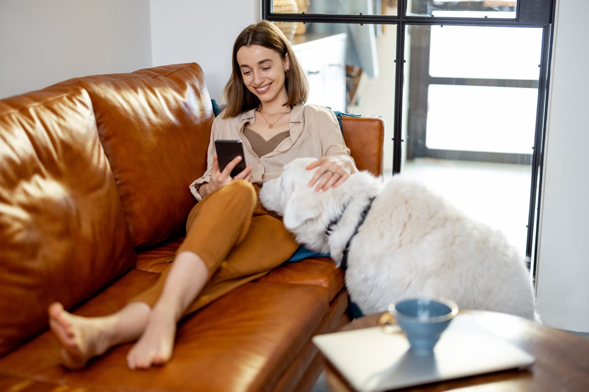 A woman relaxing on a brown leather couch, using her phone, with a white dog resting beside her.