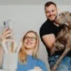 A smiling woman takes a selfie while a man holds a large gray dog in his arms indoors.