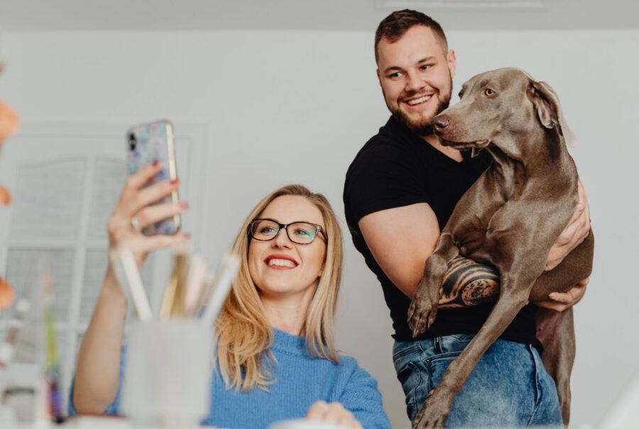 A smiling woman takes a selfie while a man holds a large gray dog in his arms indoors.