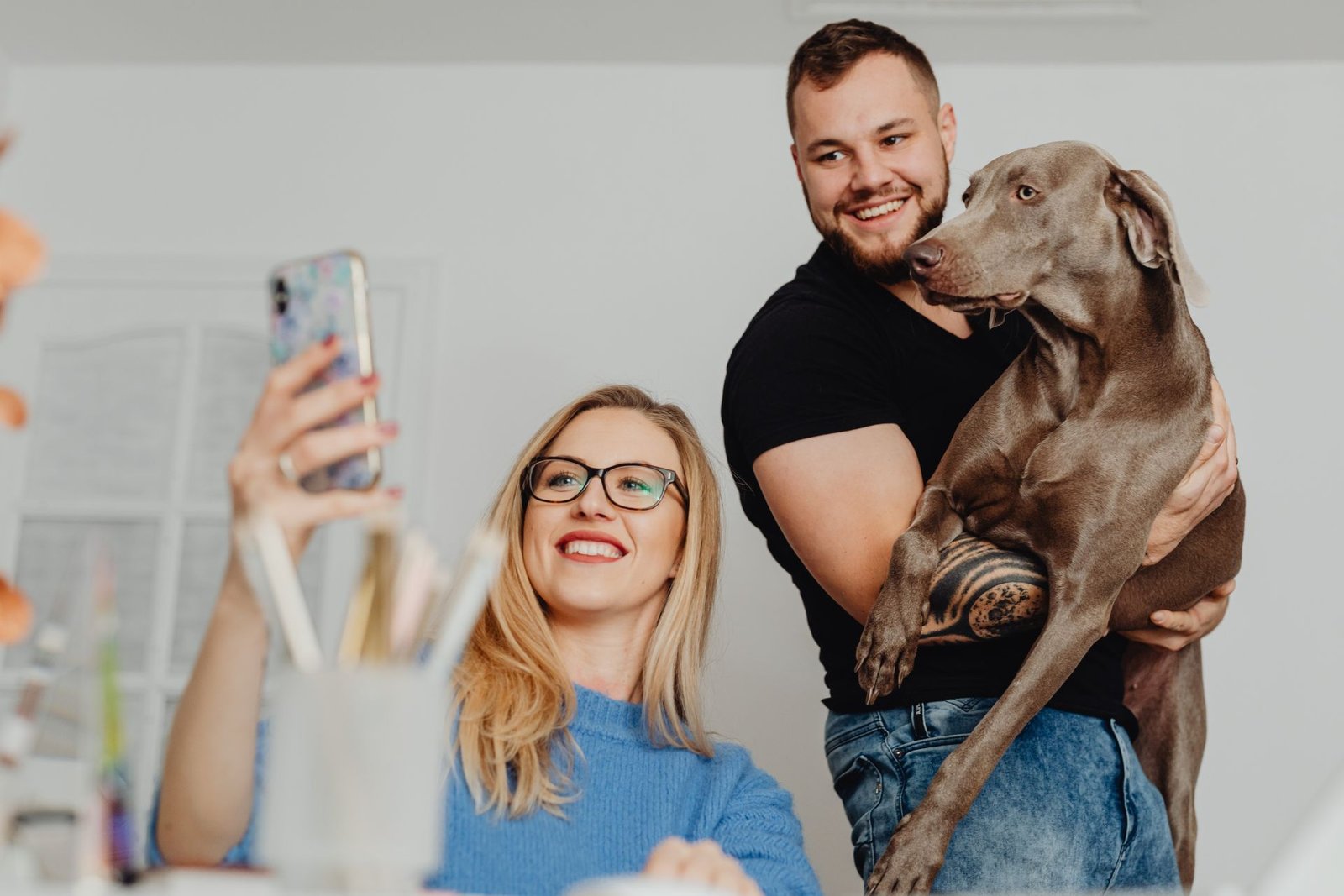 A smiling woman takes a selfie while a man holds a large gray dog in his arms indoors.
