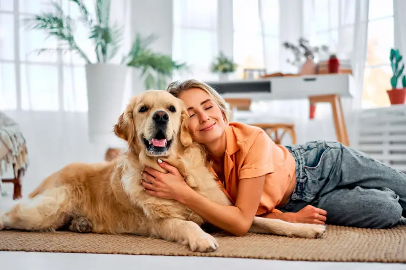 A smiling woman lies on the floor hugging a happy golden retriever in a bright, plant-filled room.